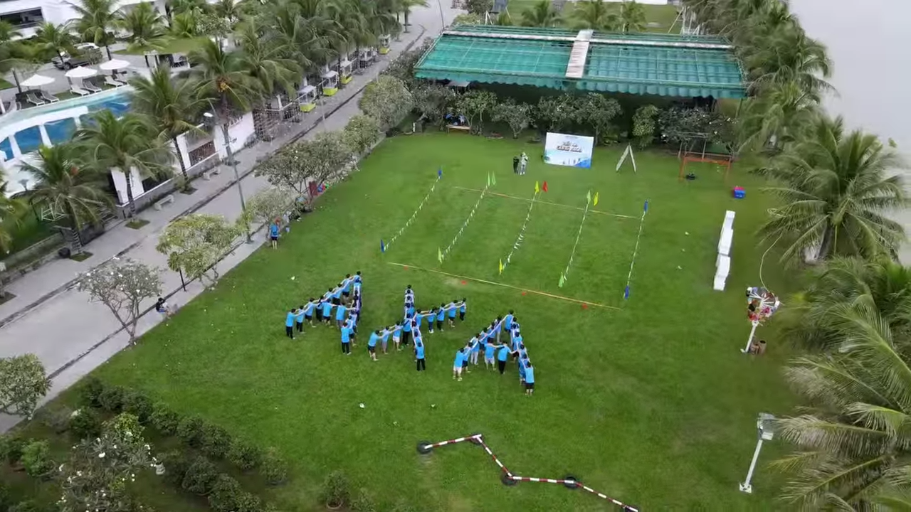 Aerial view of a lush green lawn with people in blue shirts forming the letters APA. This meticulously organized area, possibly planned with civil engineering precision, is sectioned off with markers leading to a net for a team event. Palm trees and a body of water gracefully envelop the scene.